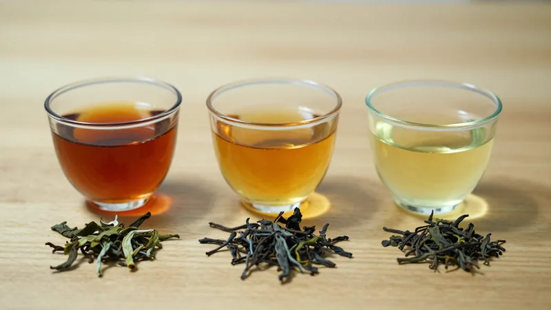 Three glass cups of brewed tea on a light oak table, one dark reddish-brown, one golden amber, one pale green-yellow, with a small pile of dry leaf in front of each