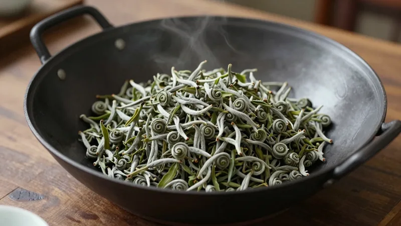 Tight, downy green spiral tea leaves in a wide wok-shaped iron pan on a wooden counter, side light