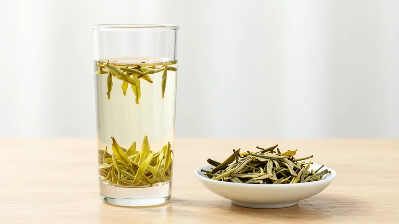 Pale yellow-green Anji Bai Cha leaves unfurling in a clear glass of hot water next to a dish of dry leaves