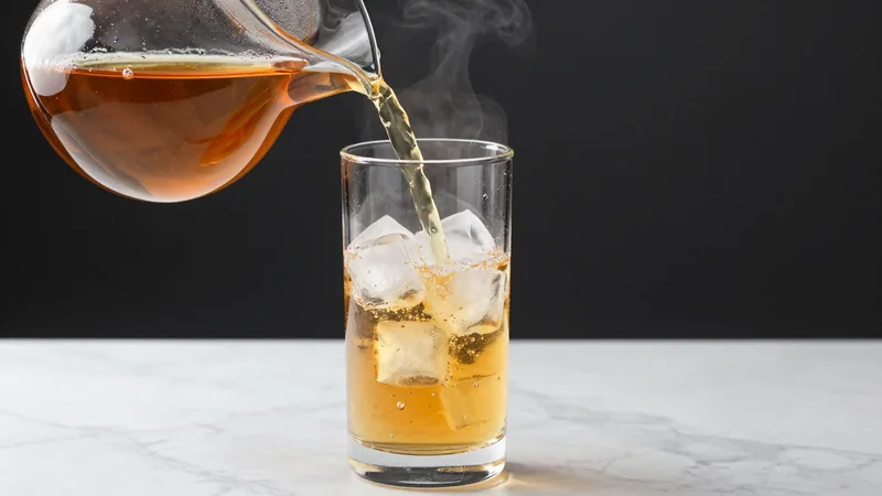 Hot amber tea being poured from a glass kettle into a tall glass pitcher packed with ice cubes on a marble counter
