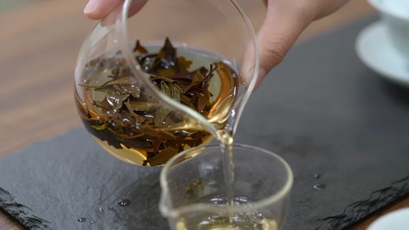 Glass gaiwan of wet dark tea leaves tilted over a glass pitcher, a thin stream of amber rinse water pouring out