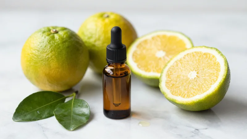 Halved green-yellow bergamot oranges on a marble counter next to a small amber glass bottle of essential oil