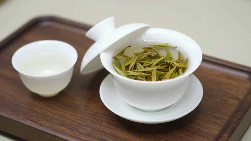 White porcelain lidded gaiwan on its saucer with the lid tilted, pale green tea leaves visible inside