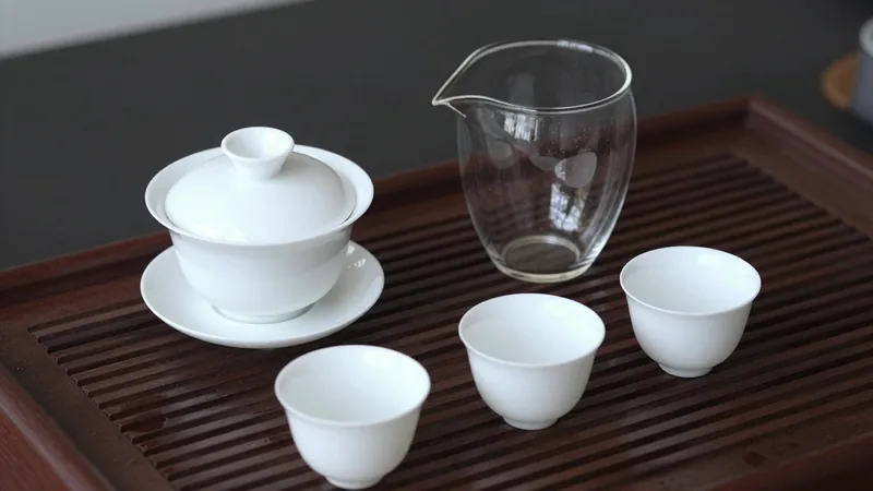 White porcelain gaiwan with its lid tilted beside a clear glass fair pitcher and three small cups on a dark wooden tea tray