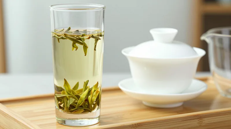 A tall clear glass of pale green tea with whole leaves floating and sinking in it, next to a lidded white porcelain gaiwan and a small glass pitcher on a bamboo tray