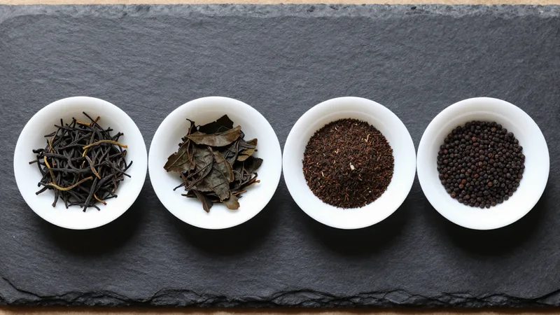 Four small white porcelain dishes of black tea in a row on a slate board: long twisted whole leaves with golden tips, broken leaf pieces, fine fannings, and round CTC pellets