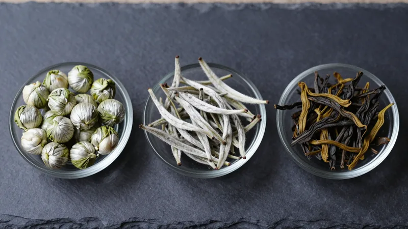 Three small glass dishes on a slate board holding rolled jasmine pearls, long silvery jasmine silver needle buds and twisted dark jasmine black tea
