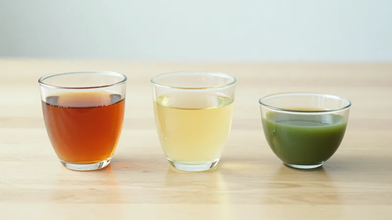 Three glass cups of tea in a row on a pale wooden table: a deep amber black tea, a pale yellow-green sencha and a small cup of thick jade-green gyokuro