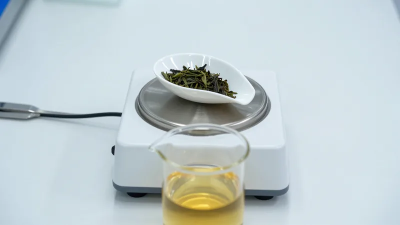 A small laboratory scale holding a white weighing boat of loose green tea leaves next to a glass beaker of brewed tea on a white lab bench