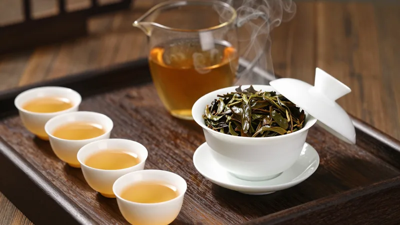 A small porcelain gaiwan with expanded wet oolong leaves beside a row of small cups of amber tea on a wooden tray