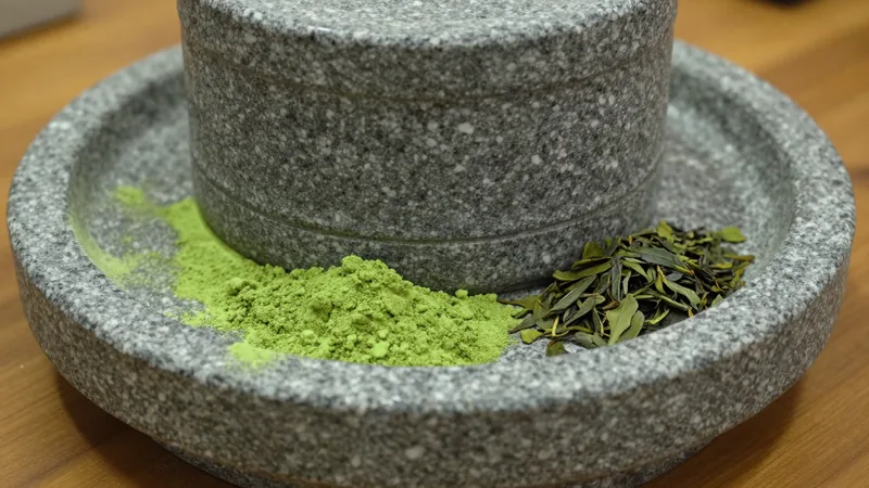 Granite hand-turned matcha mill with bright green powder collecting in its tray beside a pile of dried tencha leaf flakes