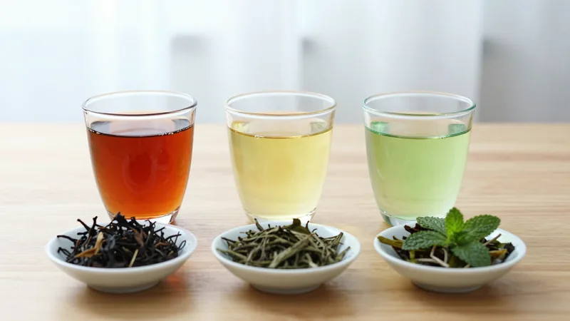 Three glass cups on a wooden table holding dark black tea, pale yellow-green tea and light green mint infusion, with loose leaves of each in small dishes in front