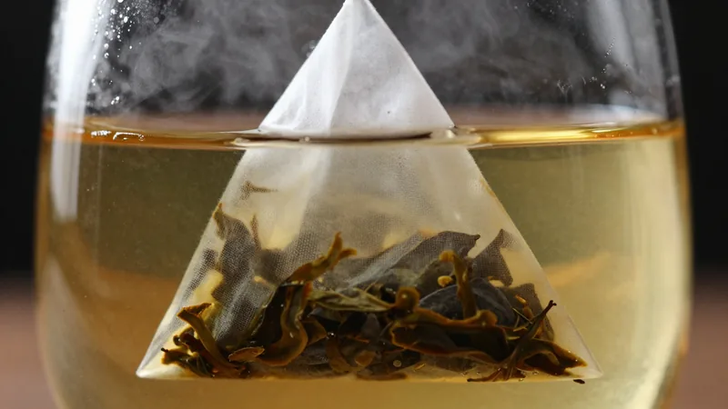 Close-up of a translucent mesh pyramid tea bag steeping in a clear glass cup of hot water, steam rising, amber tea spreading from the leaves
