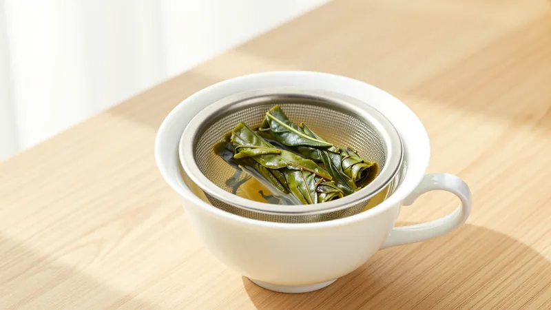 Loose oolong leaves unfurling in a stainless steel mesh infuser basket set in a white porcelain mug on a wooden table