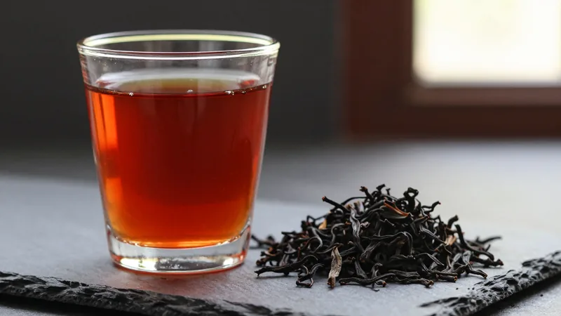 Close-up of a clear glass of bright red-orange black tea with a thin golden ring at the rim, beside a small pile of dark twisted black tea leaves on slate