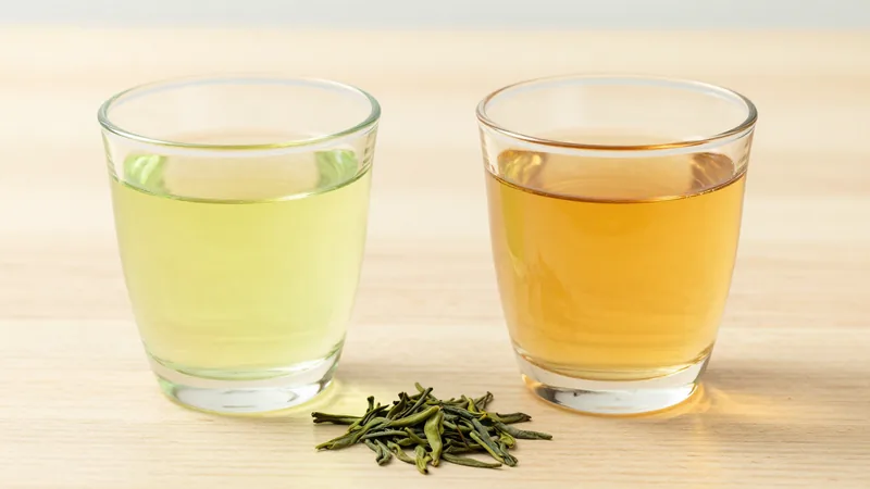 Two glasses of green tea side by side on a wooden table, one freshly brewed pale green-yellow and one darker amber after standing, with loose green leaves between them