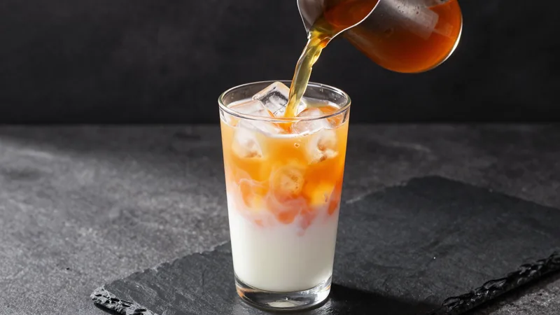 Bright orange tea being poured into a tall glass packed with ice, a layer of condensed milk visible at the bottom and pale evaporated milk swirling on top