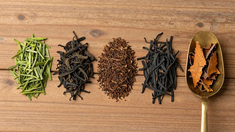 Small heaps of dry tea leaf on a wooden sorting table: bright green Japanese sencha needles, dark twisted Chinese leaf and fine brown granular black tea