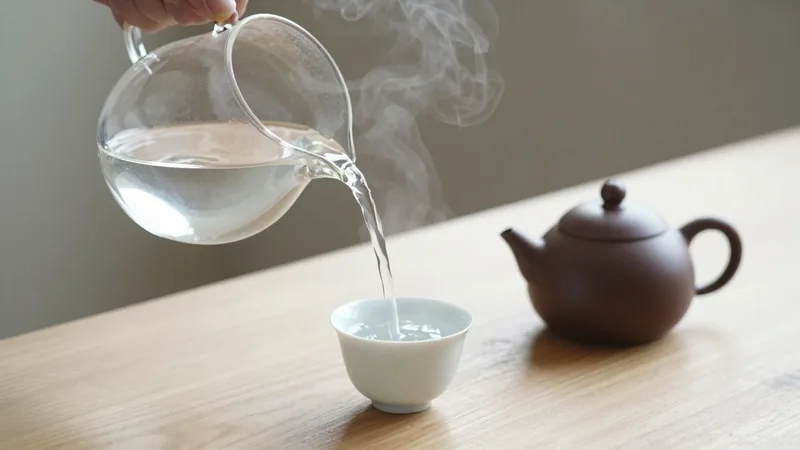 Steam rising as hot water is poured from a glass kettle into small white porcelain cups to cool before brewing, with a side-handled clay teapot nearby