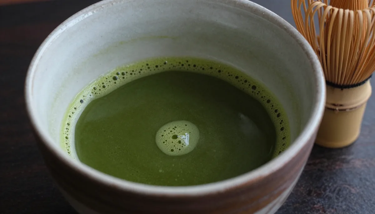 A dark raku tea bowl holding glossy, deep green thick matcha with no foam, a bamboo whisk resting on its side next to a wooden scoop